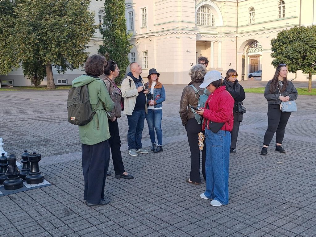 A group of about twelve project participants standing in a paved square in front of a classical white building in Vilnius; one of them uses a white cane.