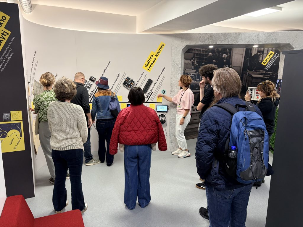 Participants gathered around an interactive exhibition wall with displays, headphones and a tablet, while a host presents the exhibits.