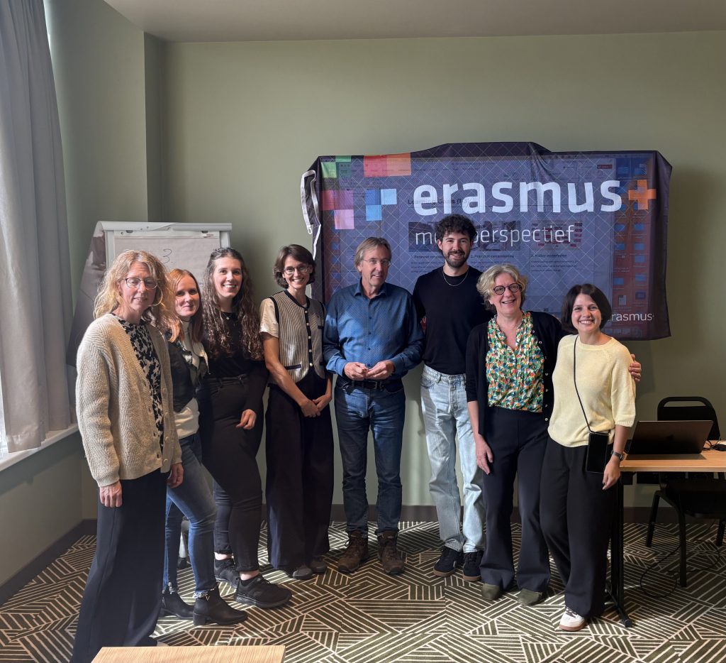Eight project team members standing side by side and smiling in front of a large Erasmus+ banner in a meeting room.