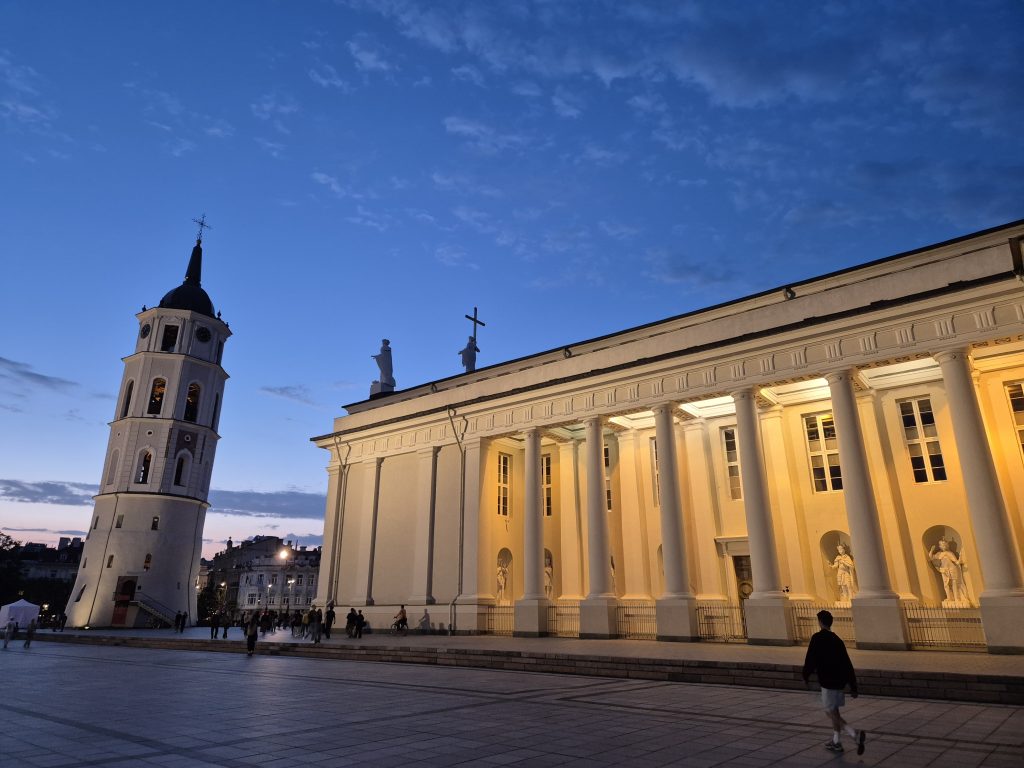 Vilnius Cathedral and its separate bell tower lit warmly against a deep blue evening sky, with a nearly empty square in the foreground.