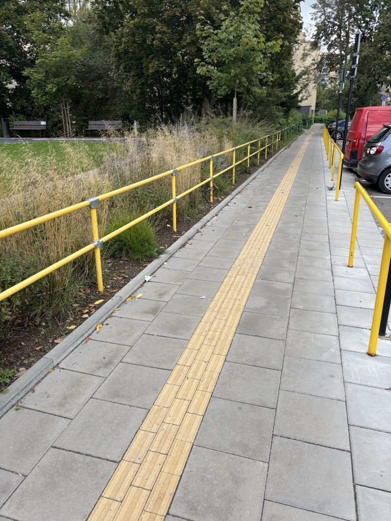 A long paved path with a yellow tactile guidance strip down the middle and bright yellow railings on both sides, with greenery on one side and parked cars on the other.