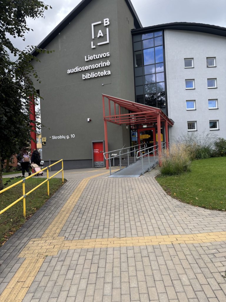 Grey library building with a white sign "Lietuvos audiosensorinė biblioteka" and a red canopy over the entrance. A ramp with metal railings leads to the door, and yellow tactile guidance strips cross the paved path in front of it.