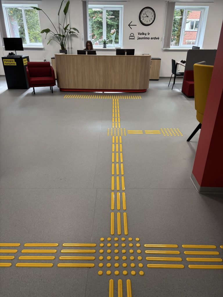 A light grey floor with yellow tactile guidance strips leading straight to the wooden reception desk; in the foreground a square of raised dots marks a crossing point.
