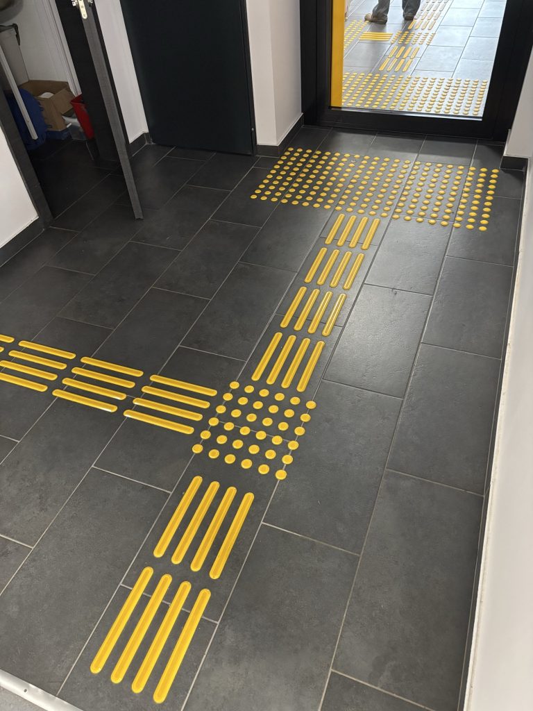 Dark grey floor tiles with yellow tactile strips: long ribbed strips meet at a square of raised dots, and a large dotted field lies in front of the doorway.