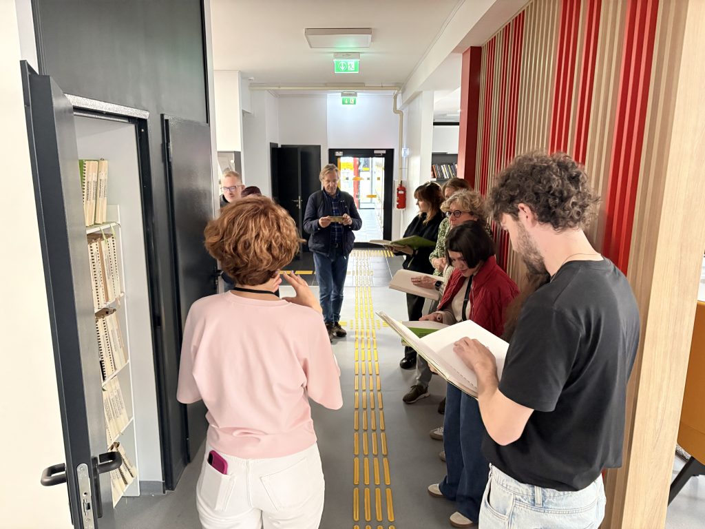 A group of visitors reads printed materials in a corridor; a clear yellow guidance path runs along the middle of the floor.