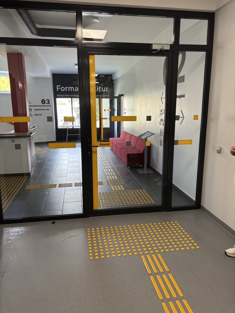 A large black-framed glass door with horizontal yellow stripes and dark squares at two heights and a yellow "Stumti" label; a dotted warning field and tactile path lead up to the door.