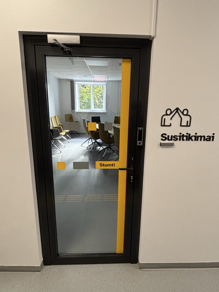 A black-framed glass door with a yellow edge and a yellow "Stumti" label, next to a raised black sign with a pictogram of two people and the word "Susitikimai" (Meetings) on a white wall.