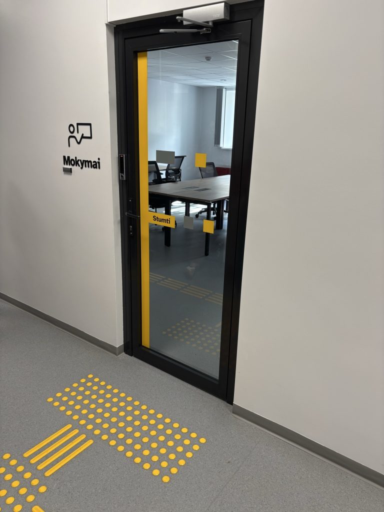 A narrow glass door with a yellow edge next to a sign with the pictogram "Mokymai" (Training); a yellow dotted warning field and guidance strips lie on the floor in front of the door.
