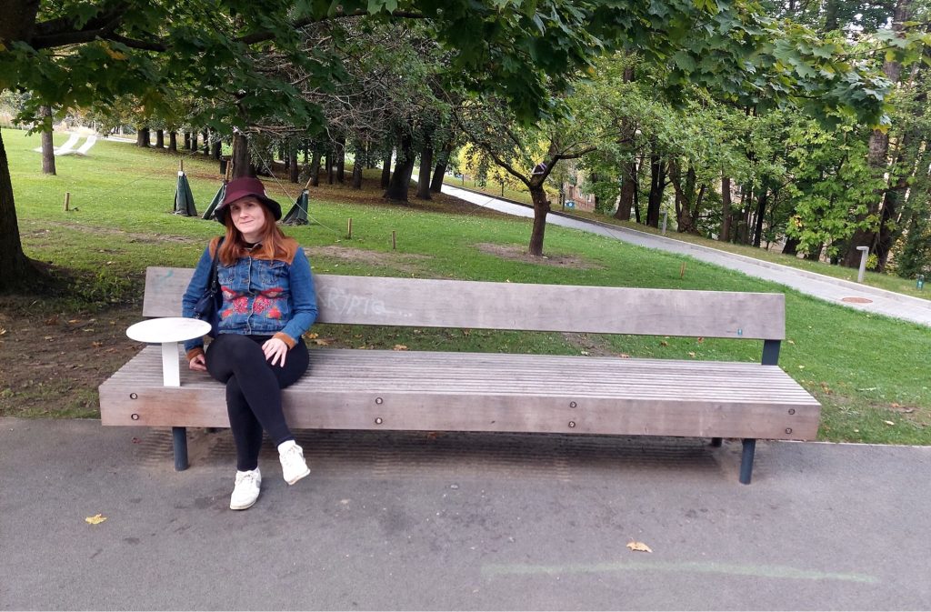A woman in a hat and denim jacket sits on a long wooden park bench with a backrest. A small round table is built into one end of the bench. Behind her is a green lawn with trees and a wide paved path.