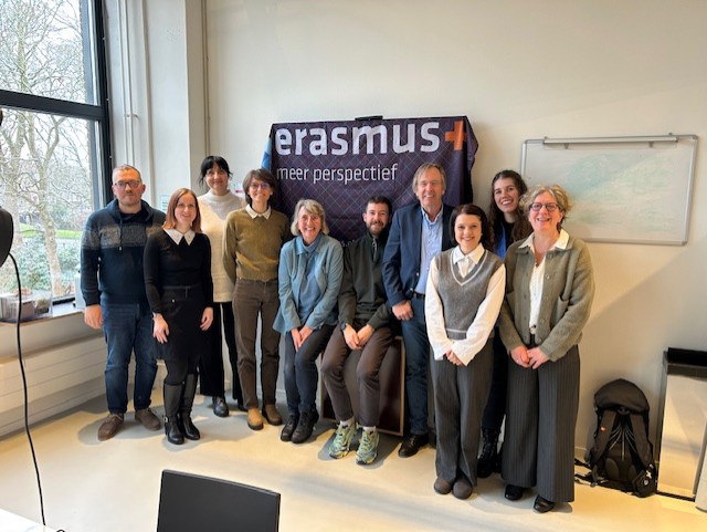 A group of ten adults standing in a row in a bright room. They are standing in front of a dark blue banner with the Erasmus+ logo and the Dutch words “meer perspectief” printed on it. Most people are smiling and facing the camera, dressed in smart-casual outfits with sweaters, shirts, and slacks; the colors are mostly neutral tones like gray, black, brown, and blue. The group includes men and women of different ages, from young adults to older adults, with a mix of hairstyles including short, medium, and long hair; hair colors range from light blond to dark brown. To the left, a large window lets in daylight, with bare tree branches and grass visible outside. On the right wall there is a whiteboard or notice board with a faint map or diagram on it, and below it on the floor is a black backpack. The floor is light-colored, and the overall atmosphere suggests an indoor meeting or workshop setting.