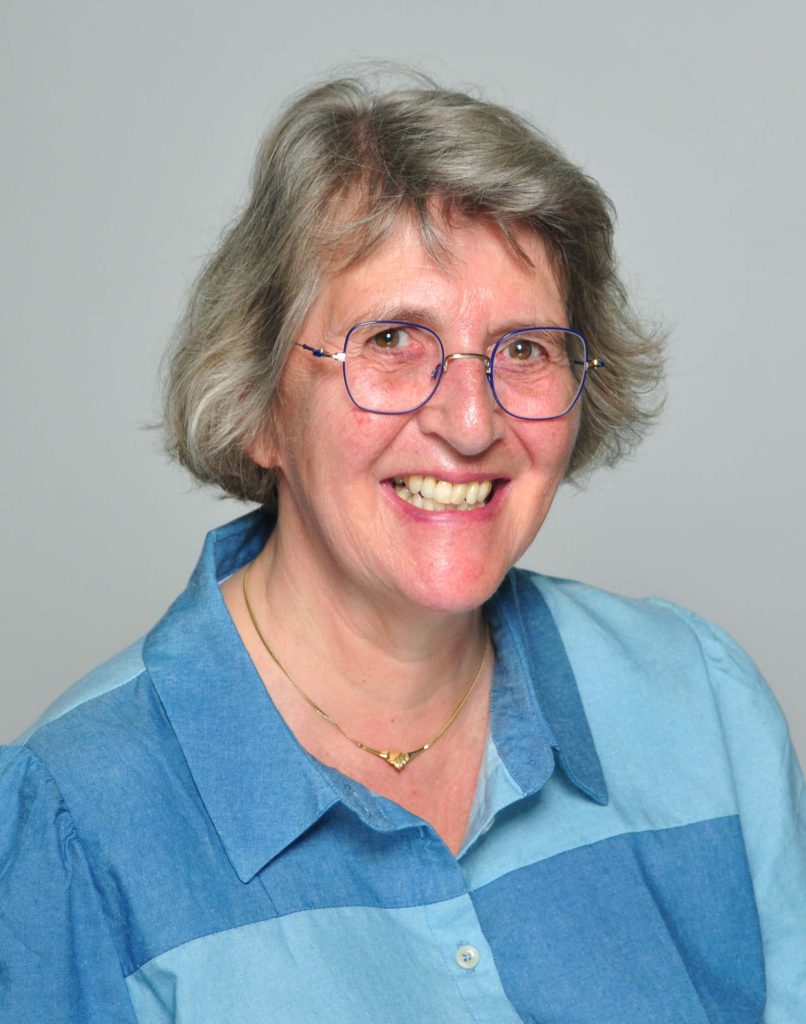 An older woman with short gray hair smiling in a studio-style portrait. She has light skin and is wearing round blue-framed glasses. Her expression is friendly, with teeth visible as she smiles. She is wearing a collared shirt made of denim-like fabric in two shades of blue arranged in large blocks. Around her neck there is a thin gold necklace with a small geometric pendant. The background is plain and light gray, with no other objects visible.